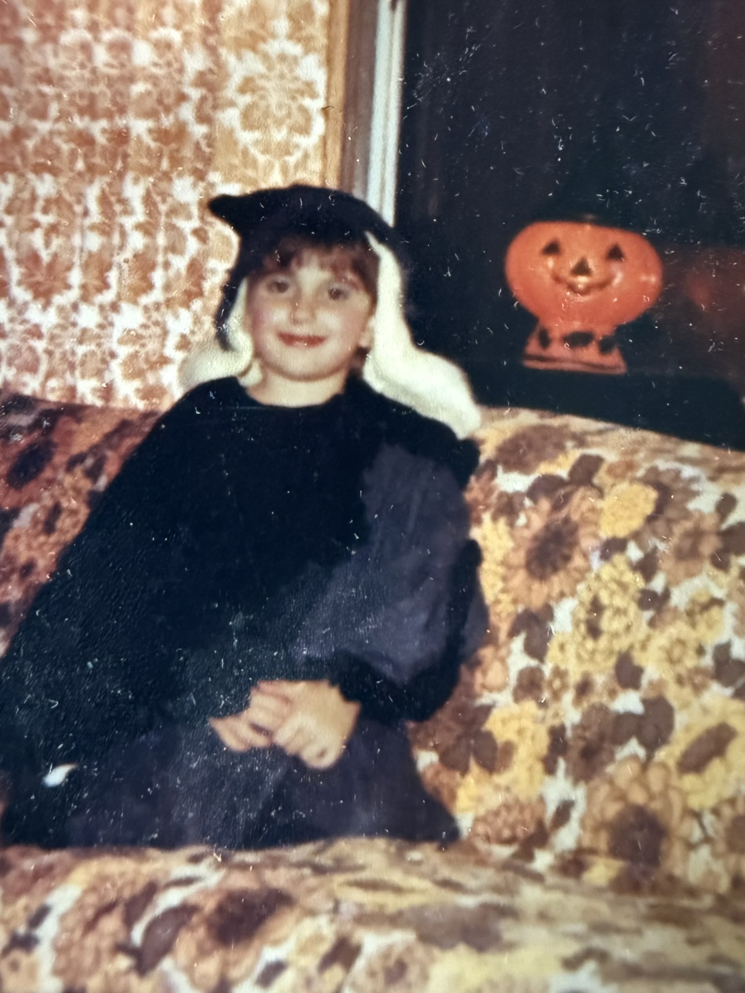 Young girl in a vintage Halloween costume sitting on a floral couch, smiling softly—early childhood photo of Rhonda Northrup.